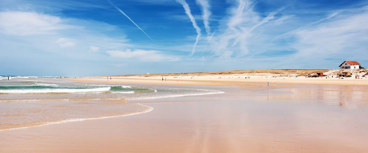 A wide sandy beach with gentle waves, a clear blue sky, and contrails.