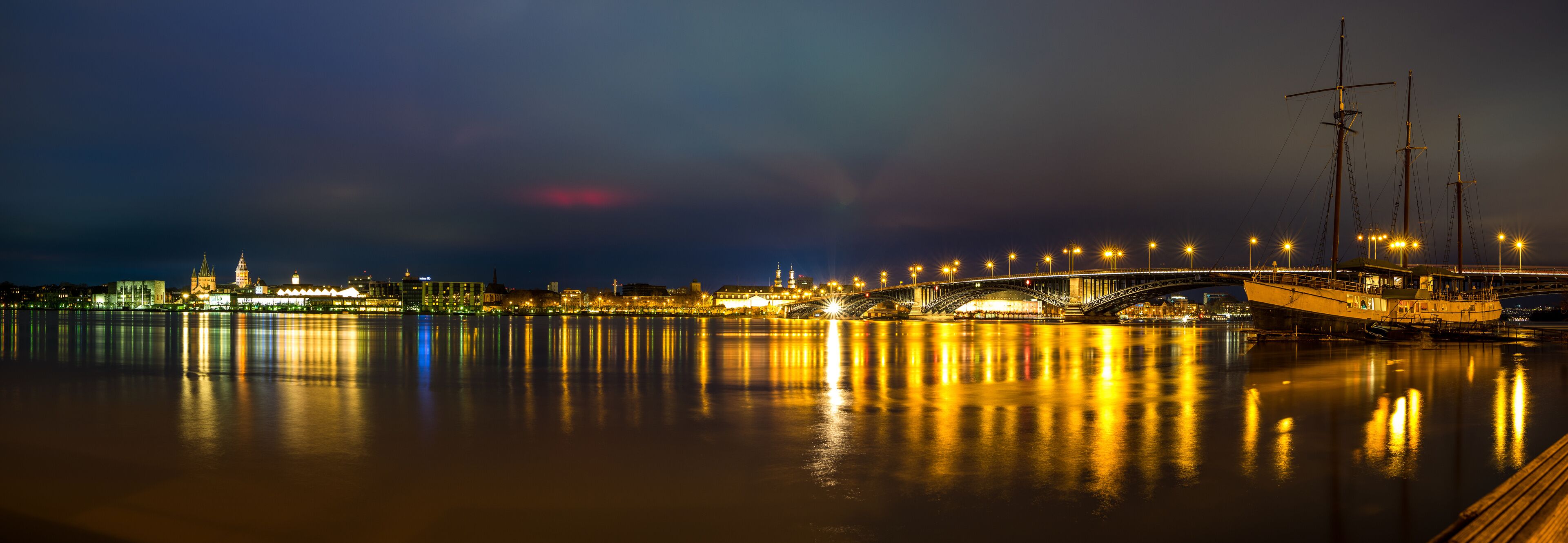A nighttime river scene with a bridge, illuminated buildings, and a boat docked on the right.