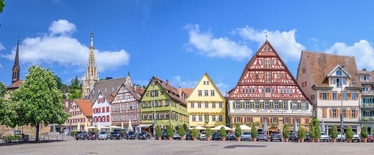 A colorful street with traditional half-timbered houses, a church spire, and parked cars.
