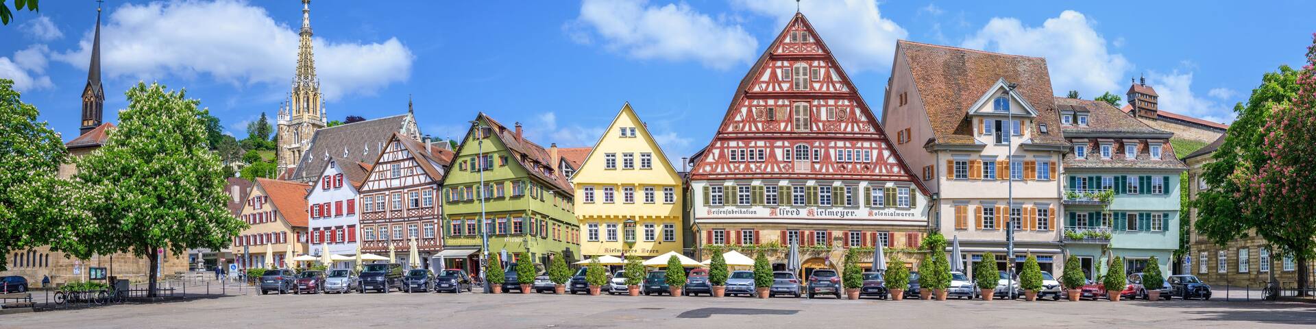 A colorful street with traditional half-timbered houses, a church spire, and parked cars.