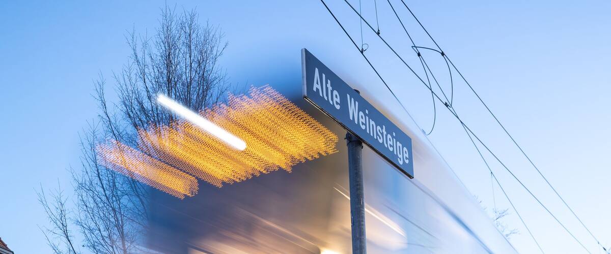 Ein fahrender Zug mit Unschärfe, ein Straßenschild mit der Aufschrift 'Alte Weinstiege' und ein klarer, blauer Himmel.