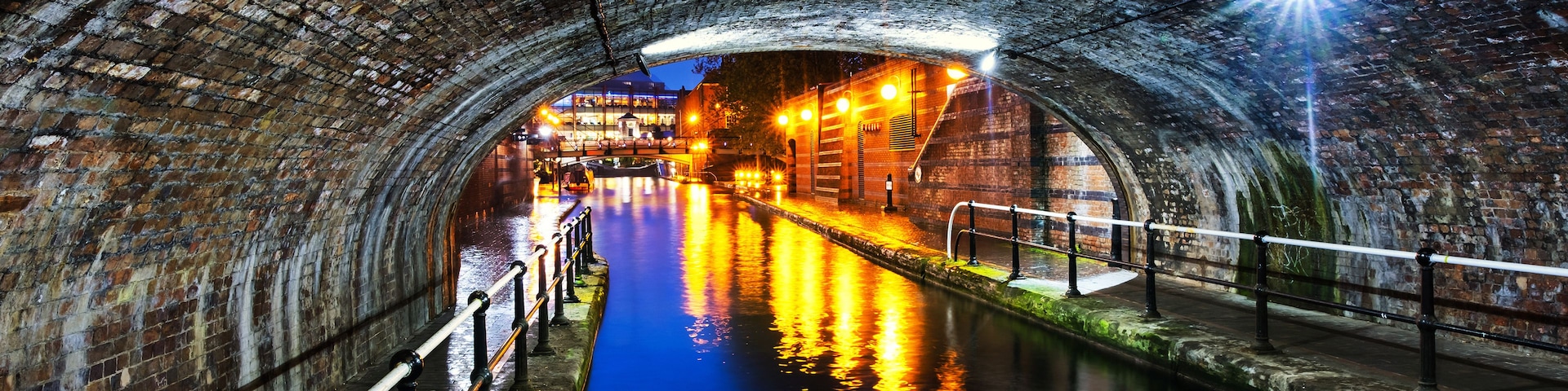 Um canal sob uma ponte de pedra com reflexos de luzes na água.