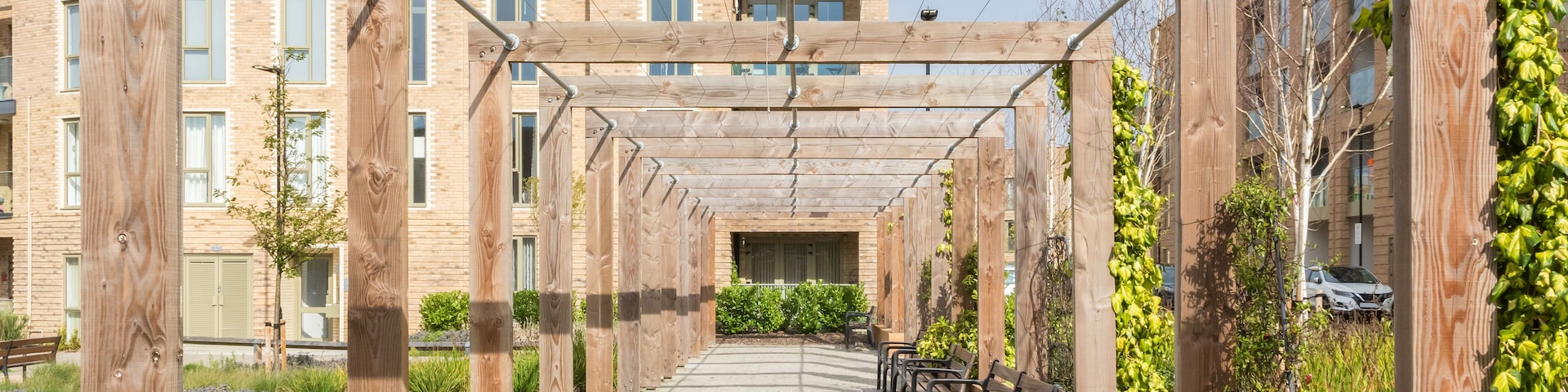 Estrutura de pérgola de madeira com bancos e plantas trepadeiras, com edifícios residenciais ao fundo.