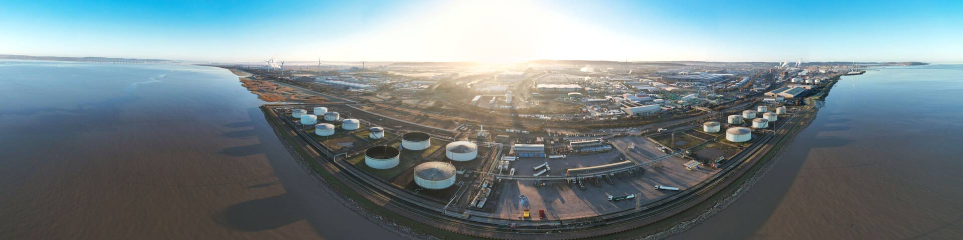 An aerial view of a large industrial complex with storage tanks, a railway, and a body of water.