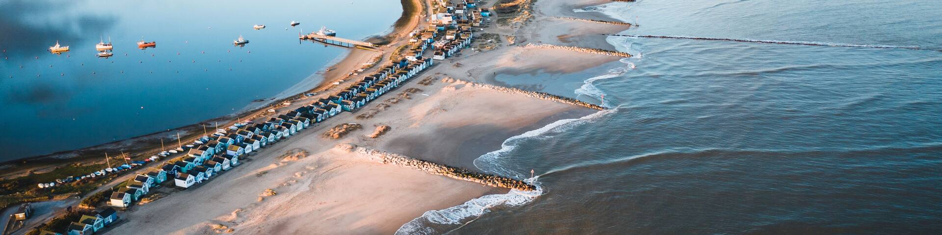 An aerial view of a coastal town with a long stretch of beach, a marina with boats, and a distant cityscape.