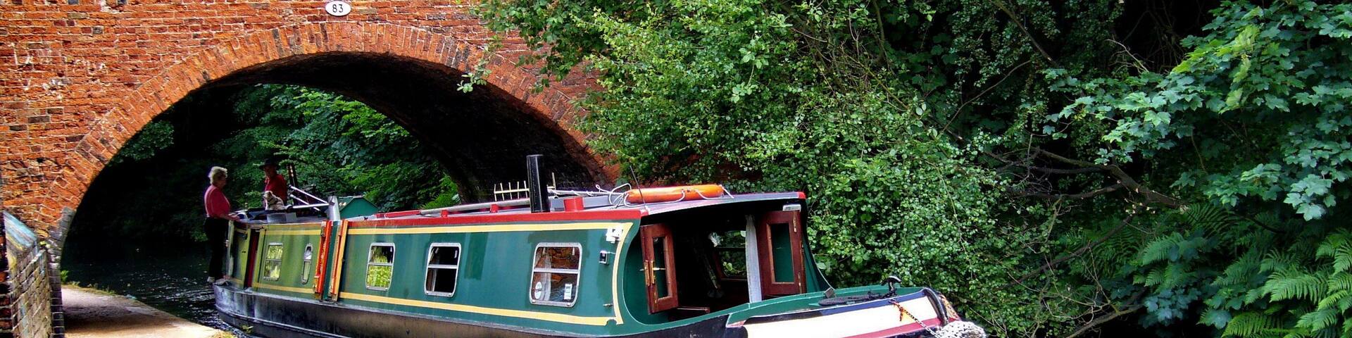 A narrowboat docked under a brick bridge with lush greenery on either side.