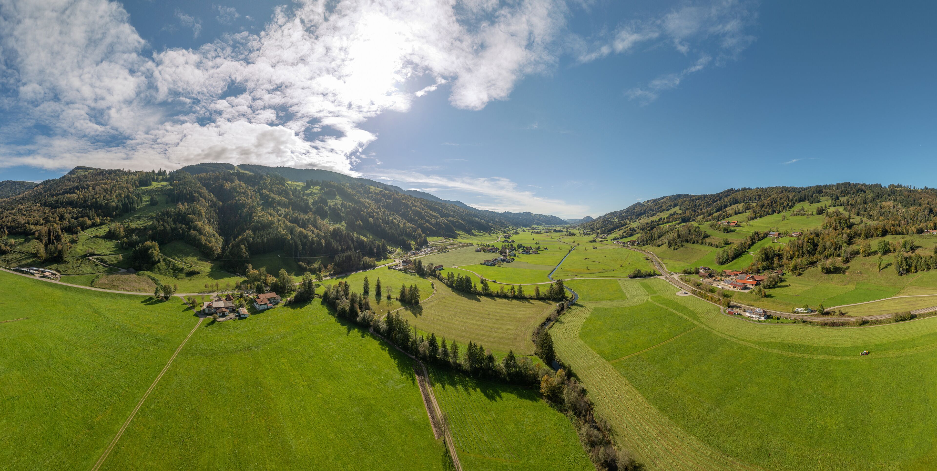 A panoramic view of a lush green valley with rolling hills, a winding road, and scattered buildings.