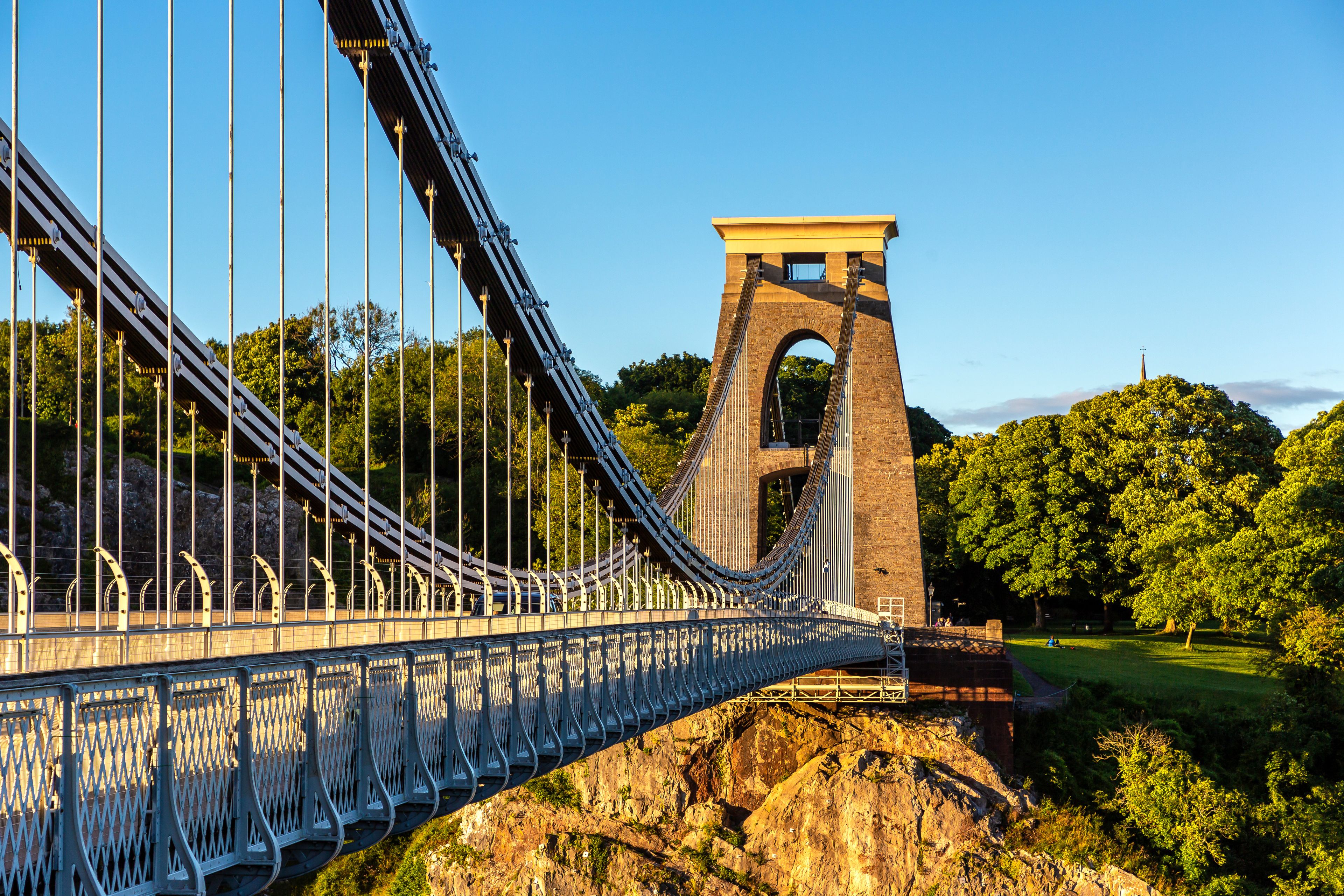 A suspension bridge with a stone tower, surrounded by lush greenery and a clear sky.