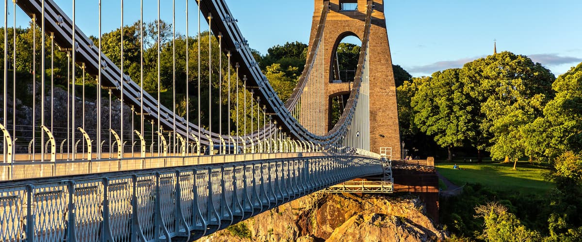 A suspension bridge with a stone tower, surrounded by lush greenery and a clear sky.