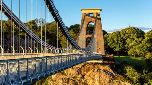A suspension bridge with a stone tower, surrounded by lush greenery and a clear sky.
