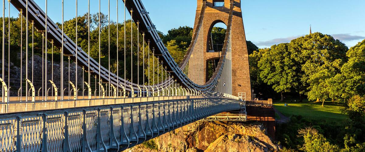 A suspension bridge with a stone tower, surrounded by lush greenery and a clear sky.