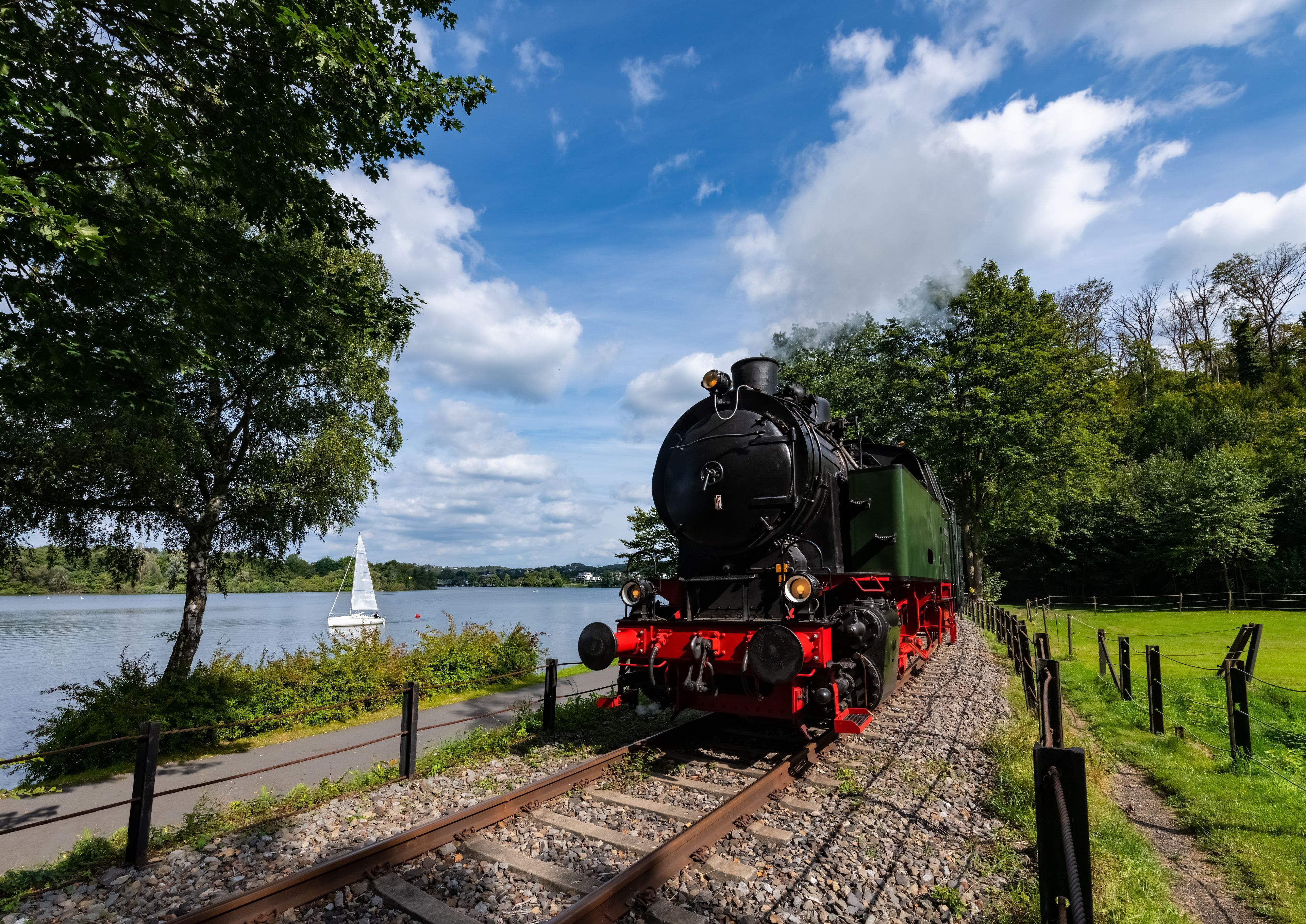 Une locomotive à vapeur sur des rails, près d’un lac, avec un voilier visible au loin.