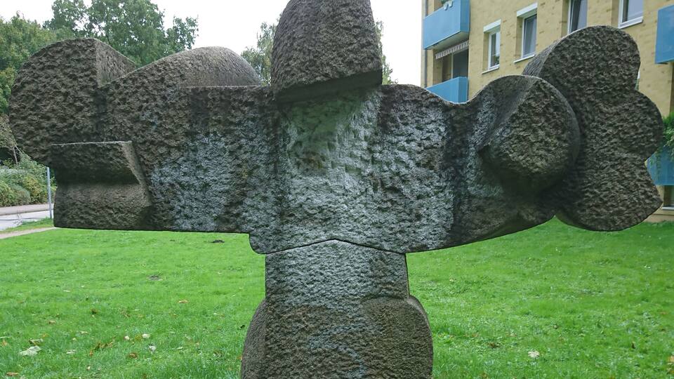 A stone sculpture of a figure with outstretched arms in a grassy area.