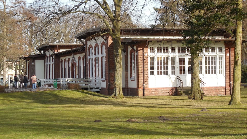 A historic building with arched windows and a brick facade, surrounded by a grassy park area with trees.