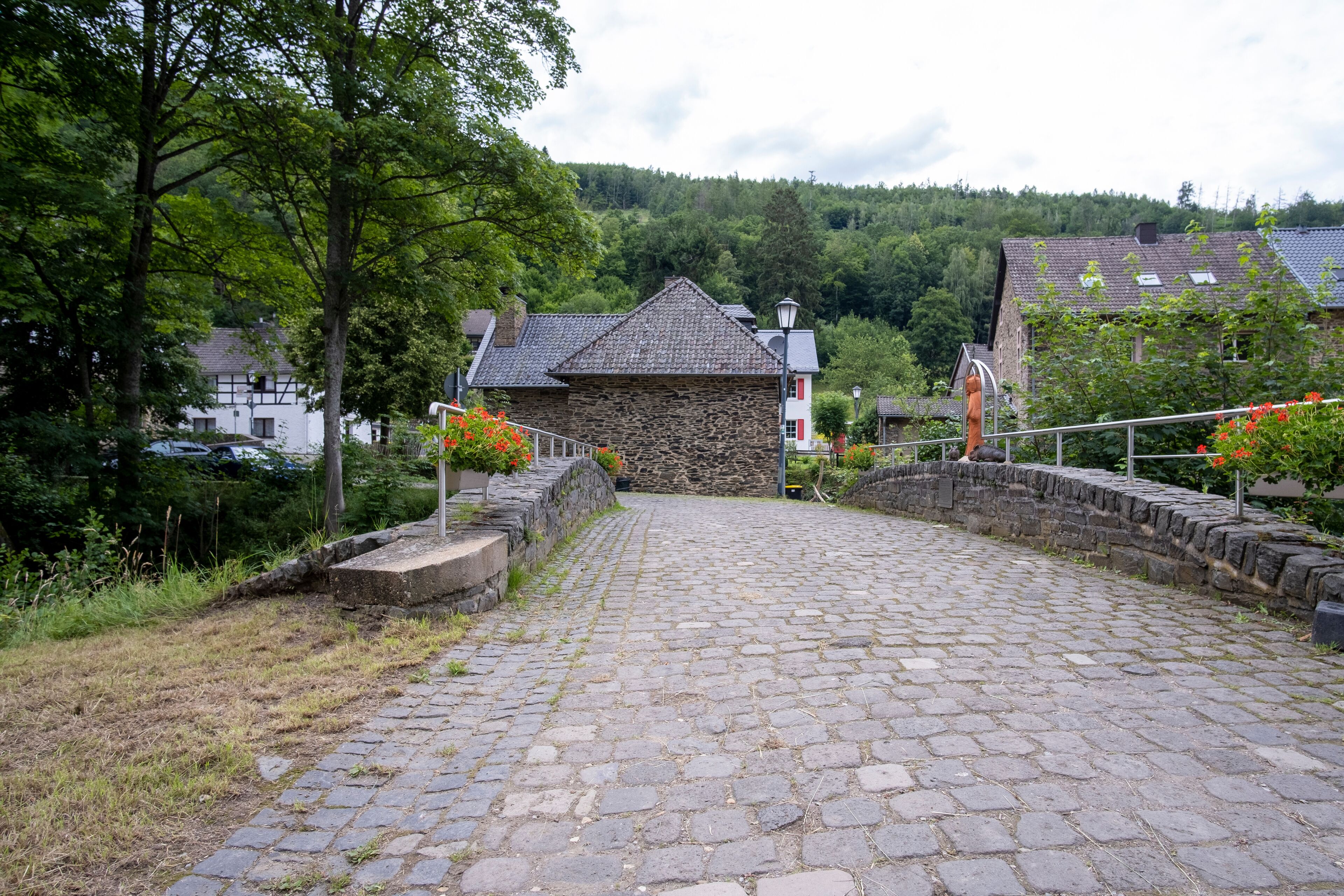A cobblestone path leading to a stone building with a sloped roof, surrounded by trees and a hillside in the background.