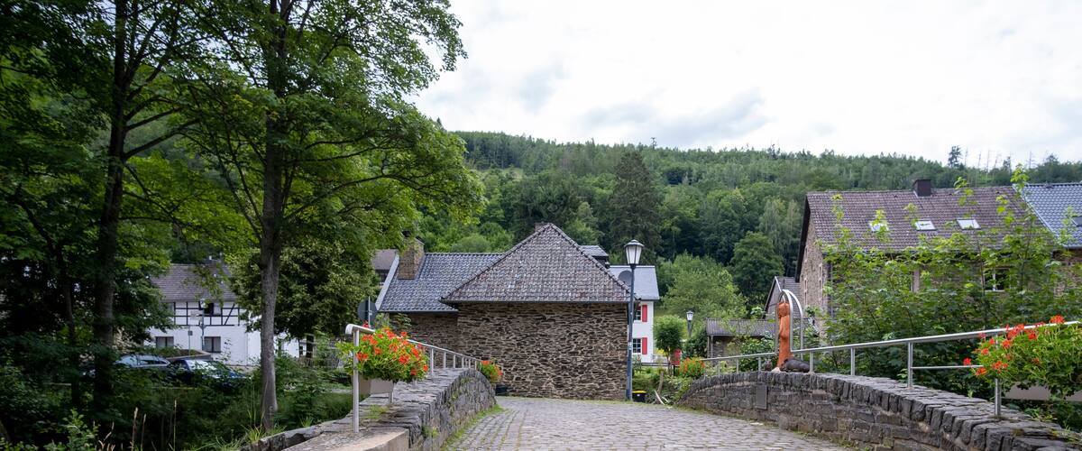 A cobblestone path leading to a stone building with a sloped roof, surrounded by trees and a hillside in the background.