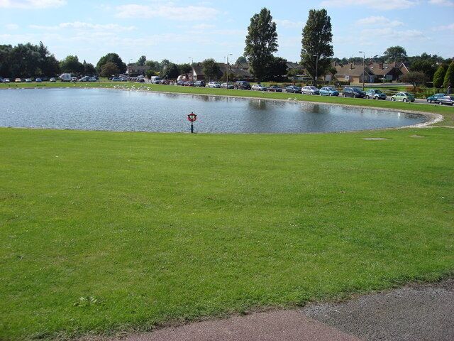 A grassy area with a pond, a parking lot, and trees in the background.