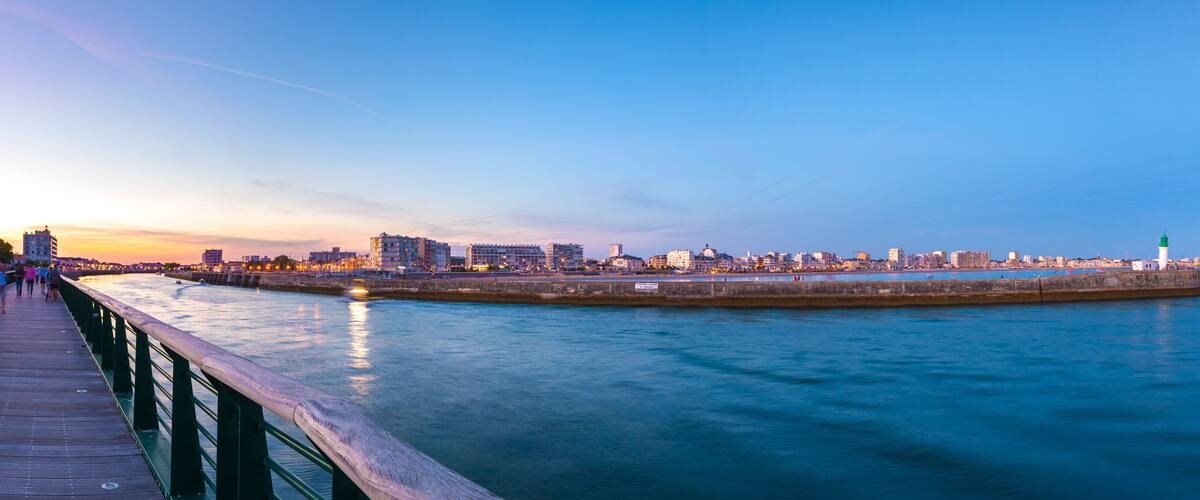 A waterfront cityscape at dusk with a pier and a lighthouse.