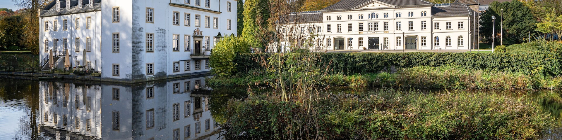 Un grand bâtiment blanc, doté de nombreuses fenêtres, dont le reflet se mire dans l’eau.
