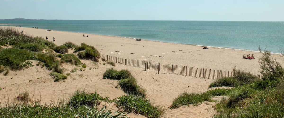 A sandy beach with a wooden fence, people walking, and the ocean in the background.