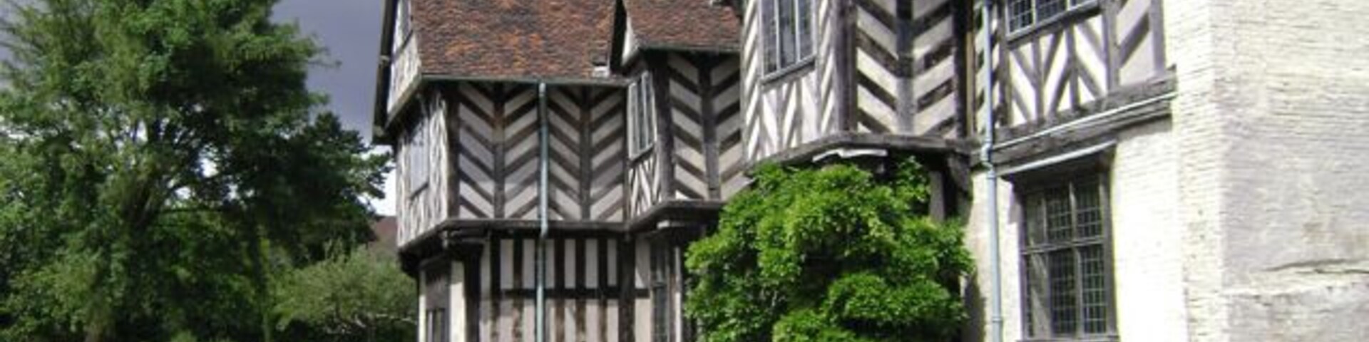 Um edifício em estilo Tudor com paredes brancas e estrutura de madeira escura, rodeado por um jardim bem cuidado e um sebe.