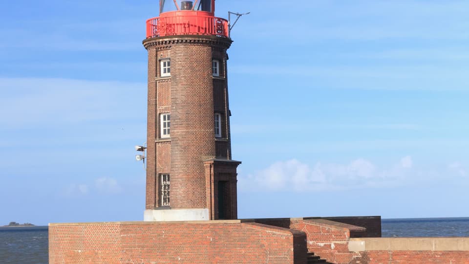 A brick lighthouse with a red dome and a weather vane on top, situated on a brick pier extending into calm waters.