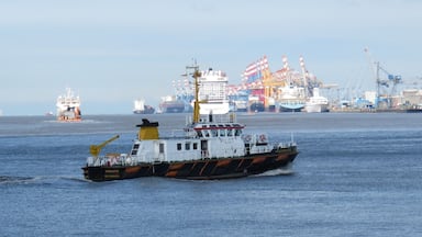 A tugboat named 'KREILOTSE' in a harbor with cargo ships and cranes in the background.