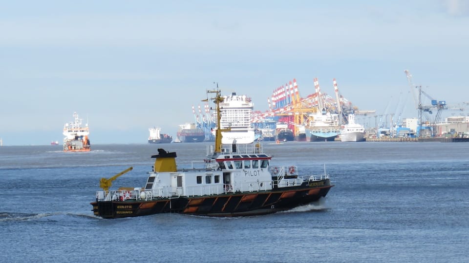 A tugboat named 'KREILOTSE' in a harbor with cargo ships and cranes in the background.