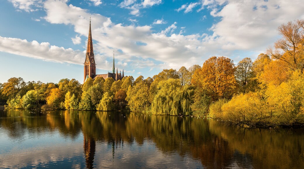 A church with a tall spire, surrounded by autumn-colored trees and a calm lake reflecting the sky.