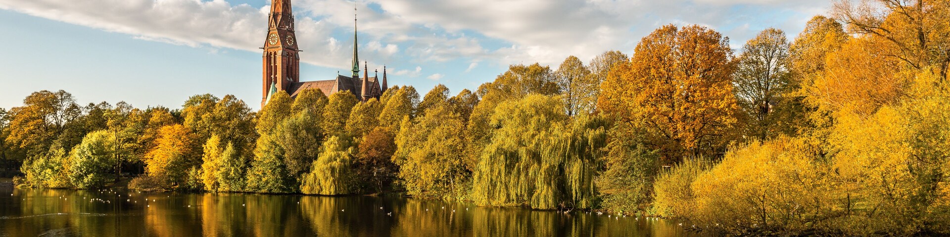 Una iglesia con una aguja alta, rodeada de árboles de colores otoñales y un lago tranquilo que refleja el cielo.