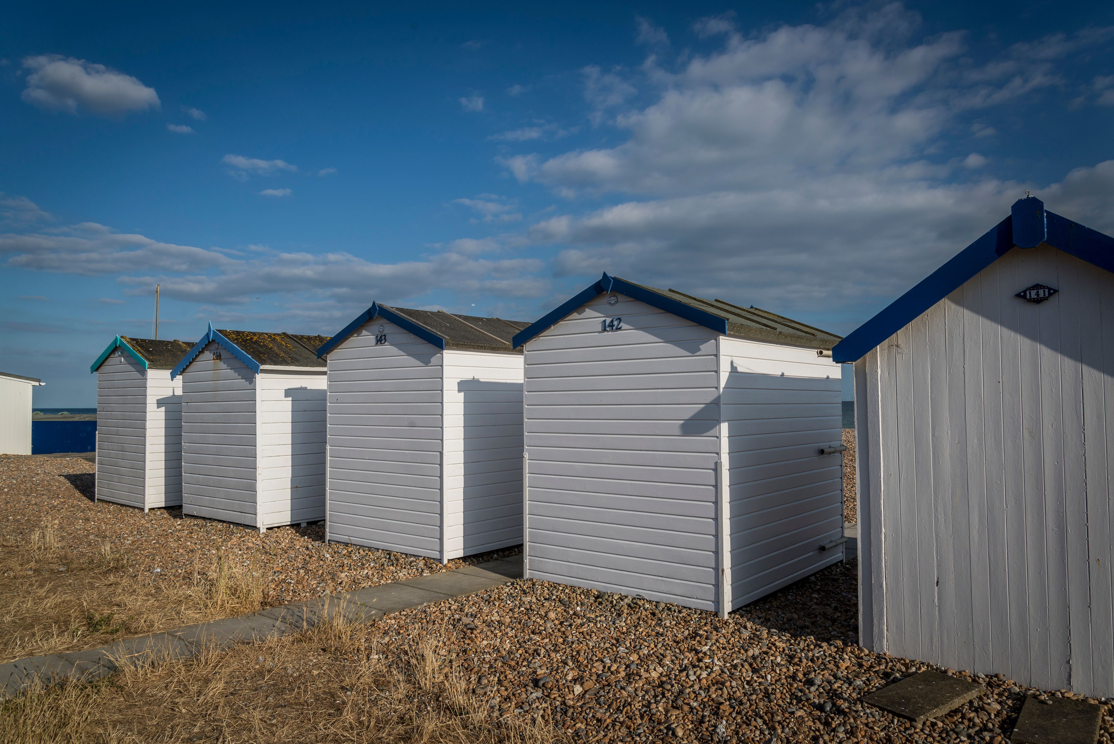 A row of white beach huts with blue trim and numbered signs, set against a clear sky and a pebble-strewn beach.