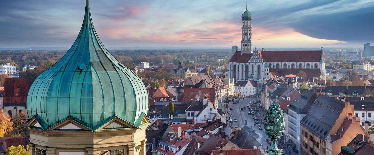 Eine Stadtansicht mit einer markanten, grün bepflanzten Kirchendome, einer historischen Kirche mit hohem Turm und einem dichten Stadtgebiet mit verschiedenen Gebäuden.