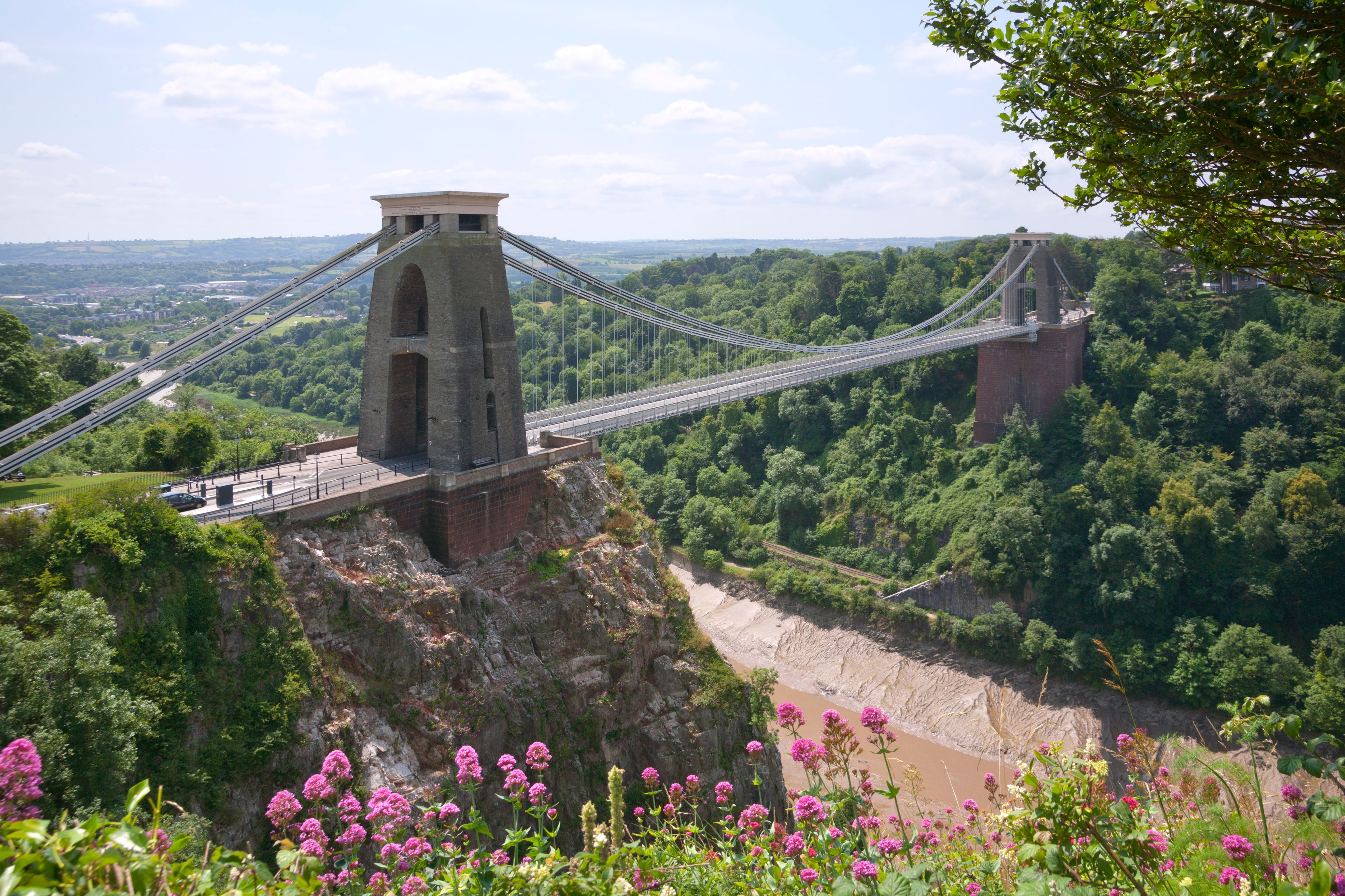 A suspension bridge with a tower, surrounded by lush greenery and a river below.