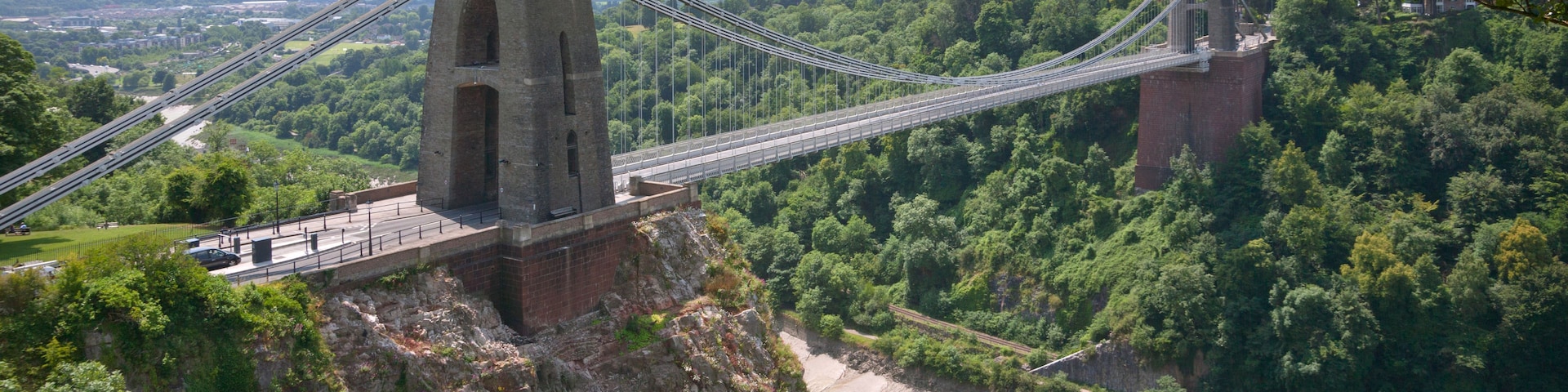 Uma ponte suspensa com uma torre, rodeada por vegetação exuberante e um rio abaixo.