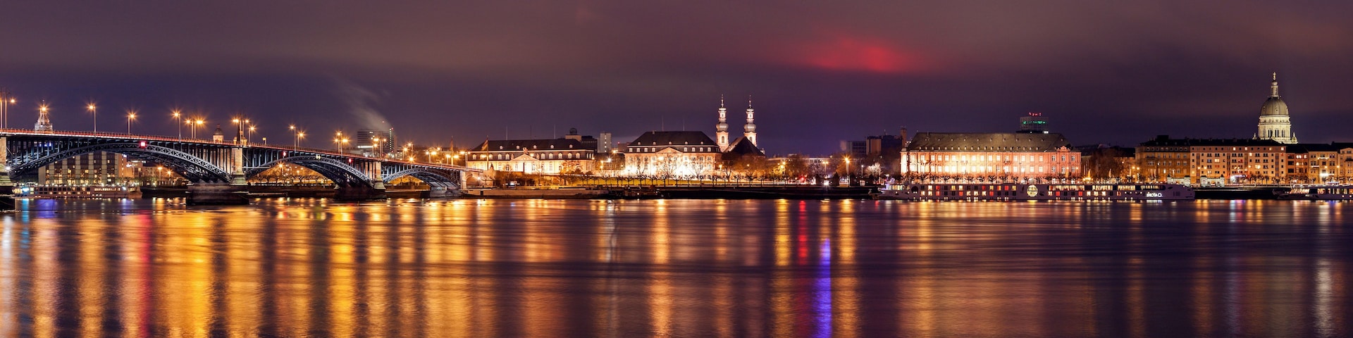 Paisagem urbana noturna com uma ponte, edifícios iluminados e um rio refletindo luzes.