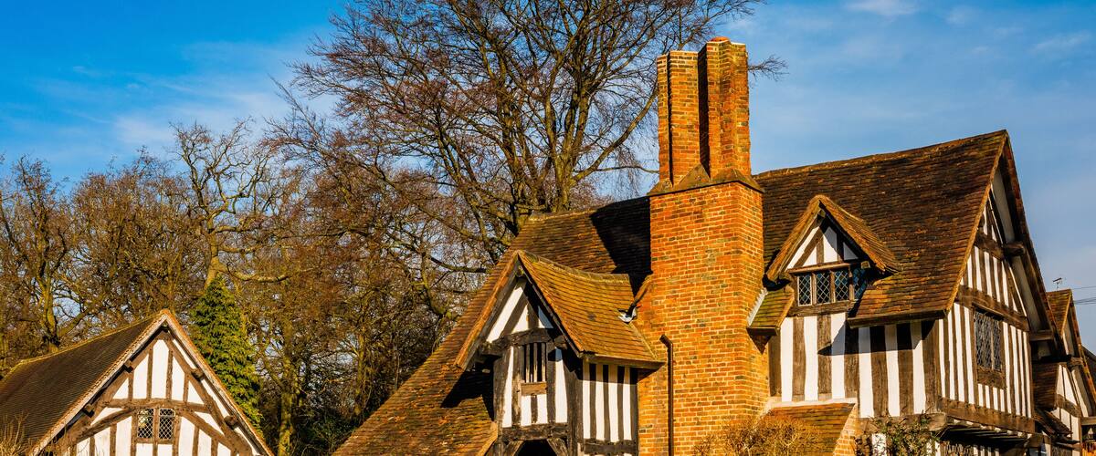 A traditional Tudor-style house with a prominent brick chimney, surrounded by a well-maintained hedge and mature trees.
