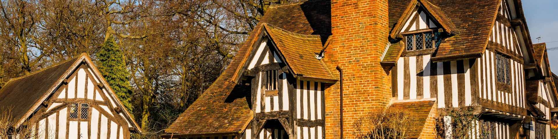Uma casa tradicional em estilo Tudor, com chaminé de tijolos destacada, rodeada por um conjunto de sebes bem cuidadas e árvores maduras.