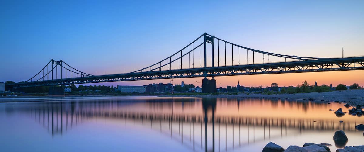 Eine Hängebrücke im Abendlicht mit einem ruhigen Fluss, der ihre Struktur spiegelt.