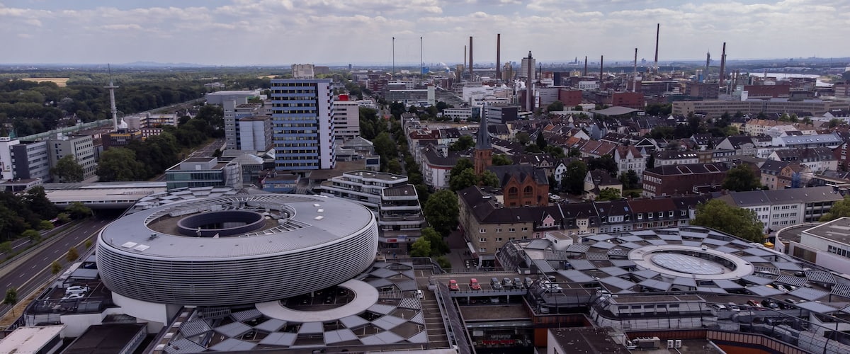 Eine Stadtansicht mit einem großen, runden Gebäude, Industrie-Schornsteinen und einer Mischung aus moderner und traditioneller Architektur.