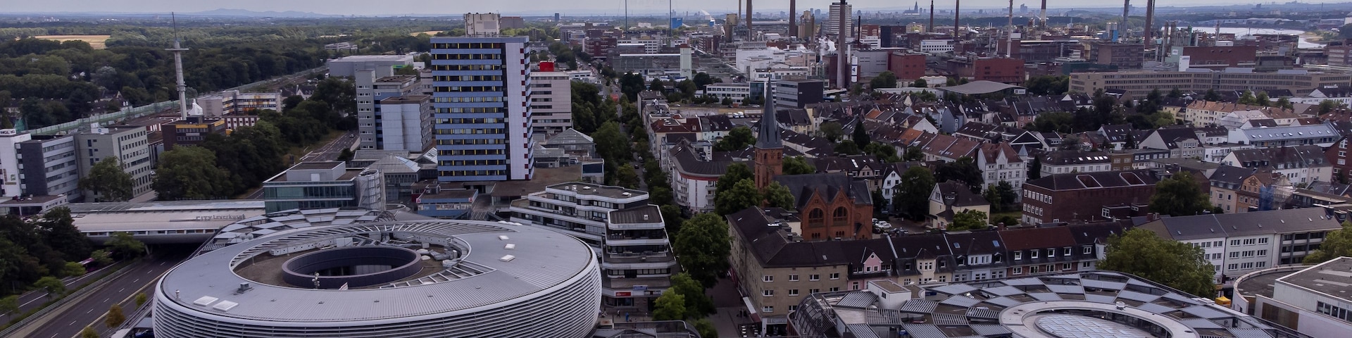 Un paisaje urbano con un edificio circular grande, chimeneas industriales y una mezcla de arquitectura moderna y tradicional.