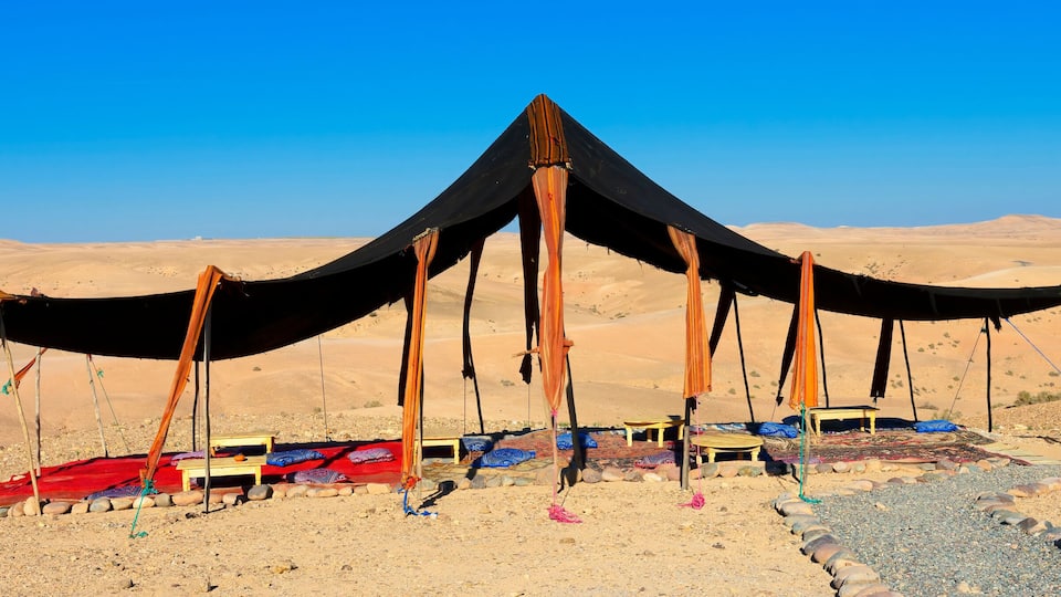 A desert tent with a black canopy and wooden poles, surrounded by colorful rugs and stones.