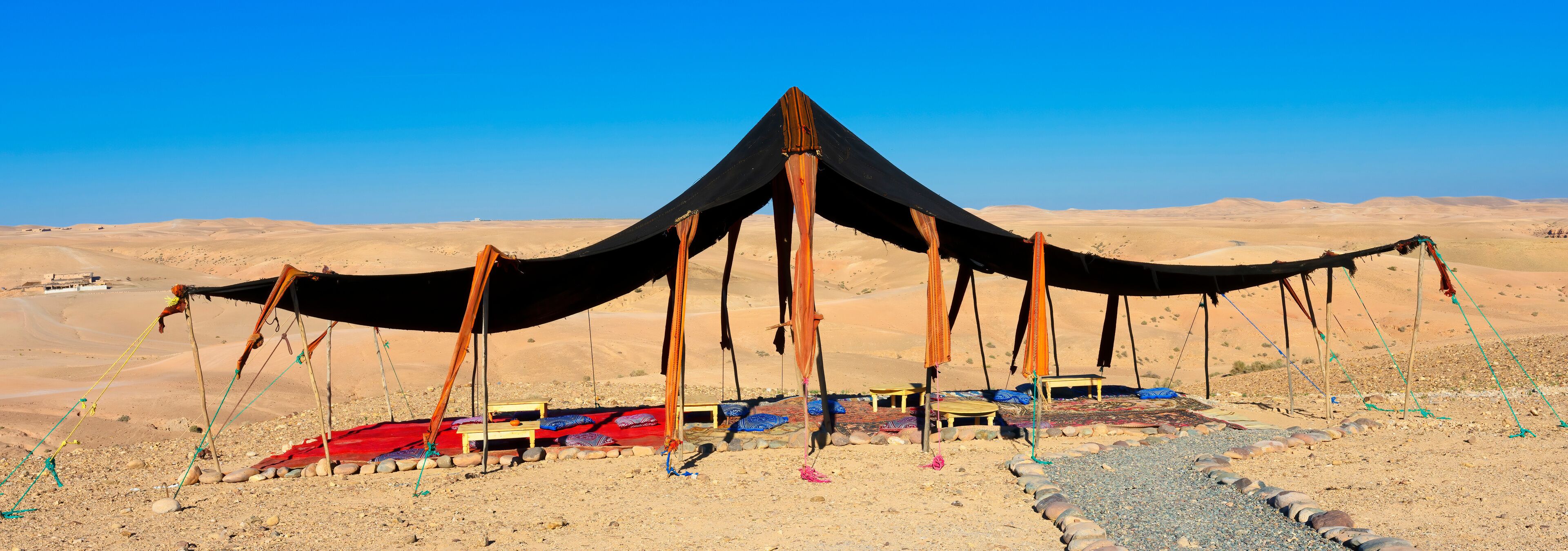 A desert tent with a black canopy and wooden poles, surrounded by colorful rugs and stones.