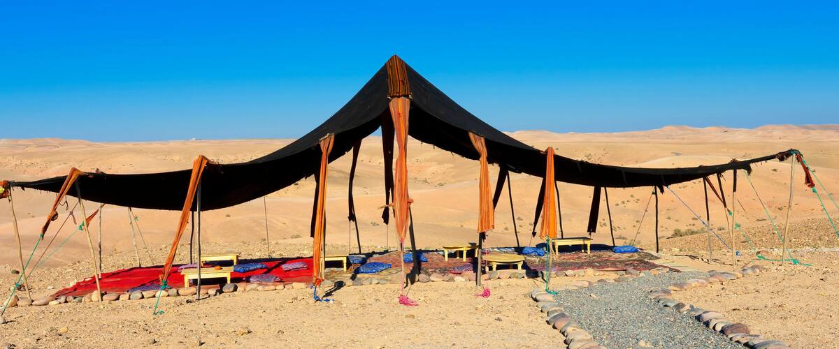 A desert tent with a black canopy and wooden poles, surrounded by colorful rugs and stones.