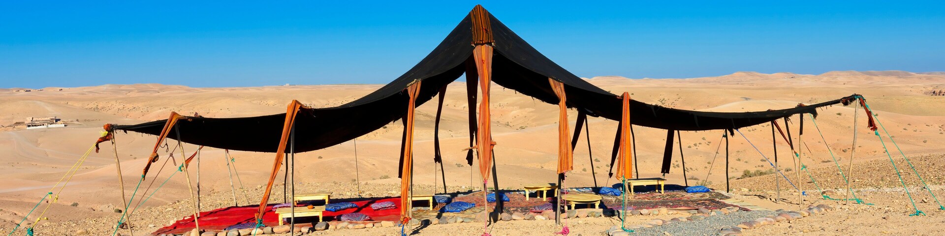 A desert tent with a black canopy and wooden poles, surrounded by colorful rugs and stones.