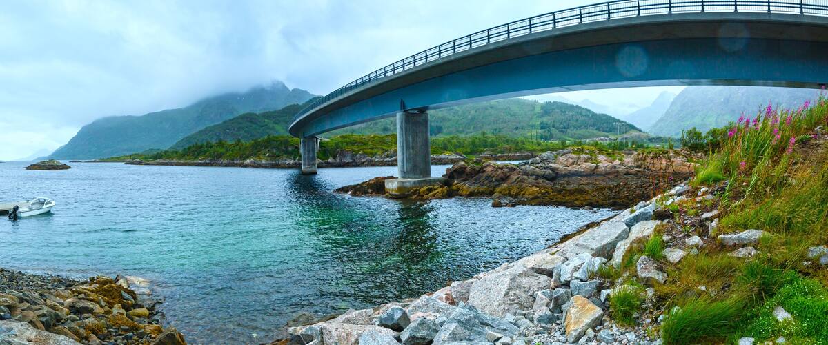 A bridge over a calm river with rocky shores and mountains in the background.