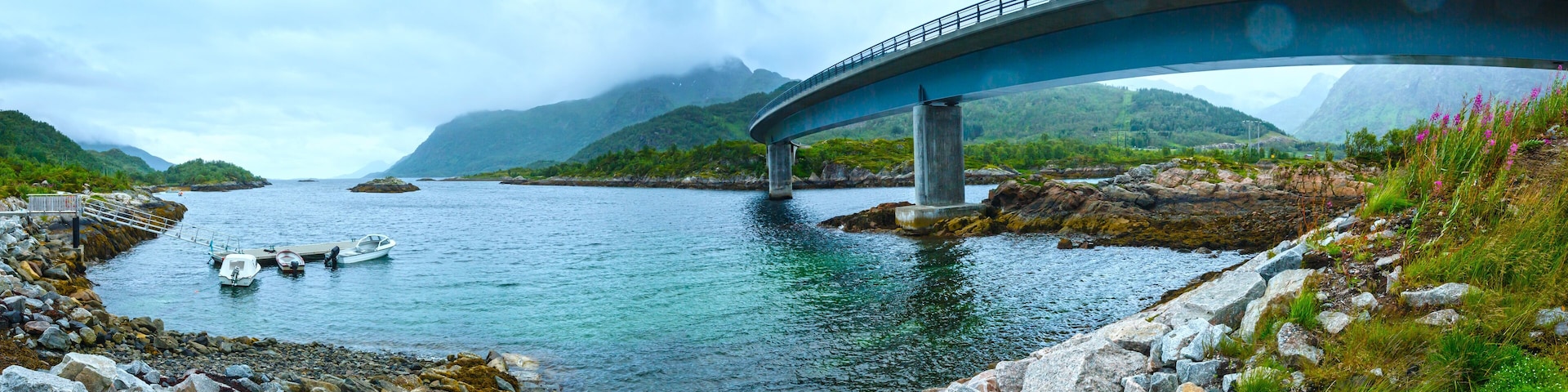 A bridge over a calm river with rocky shores and mountains in the background.