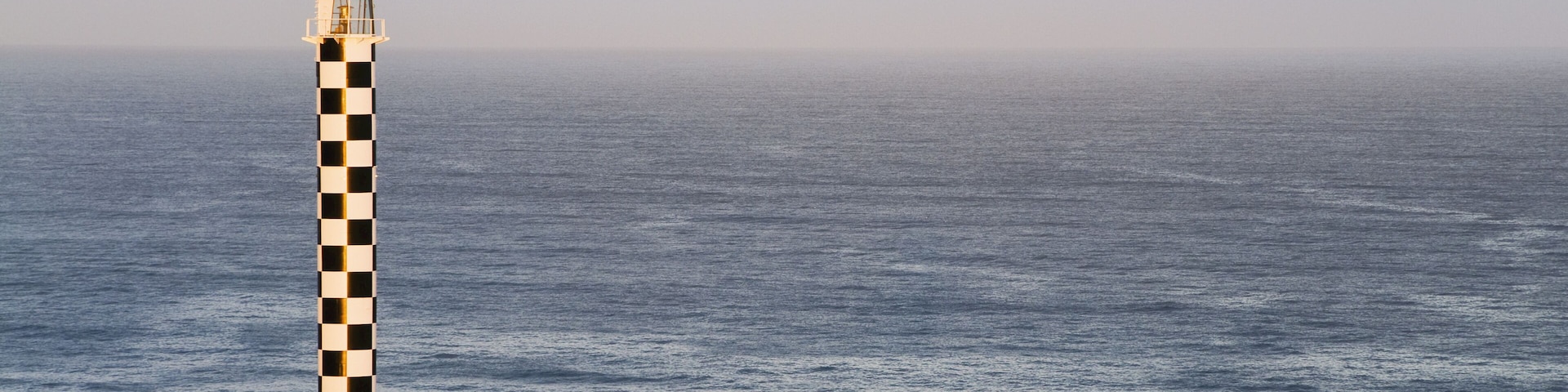 A lighthouse with a checkered pattern, a calm sea, and residential buildings with sloped roofs.