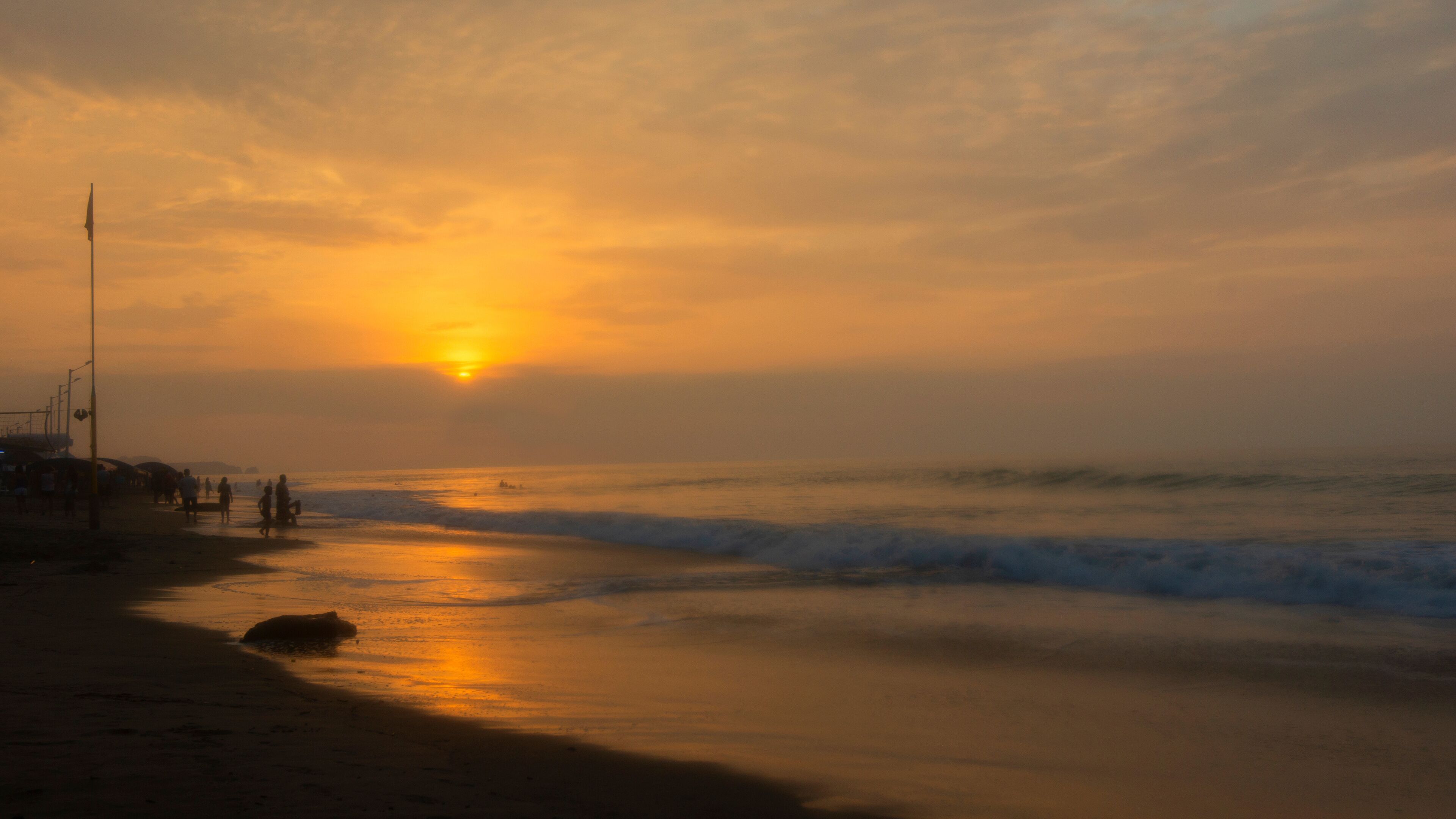A beach at sunset with the sun low on the horizon, waves gently lapping the shore, and a few people walking along the beach.