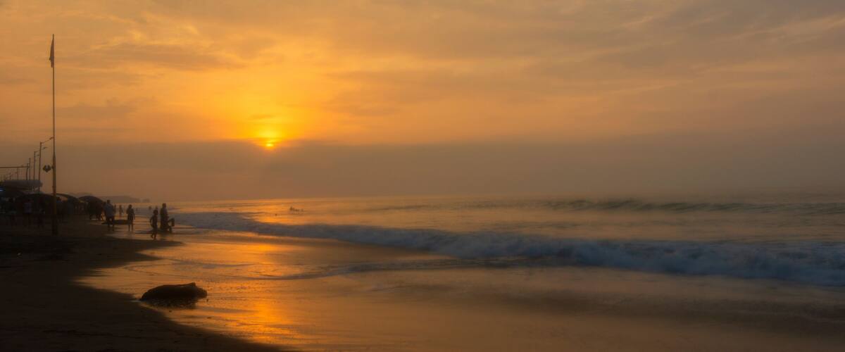 A beach at sunset with the sun low on the horizon, waves gently lapping the shore, and a few people walking along the beach.