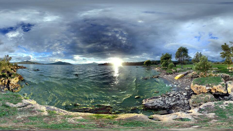 A panoramic view of a lake with clear water, surrounded by rocky shores and trees, under a cloudy sky.